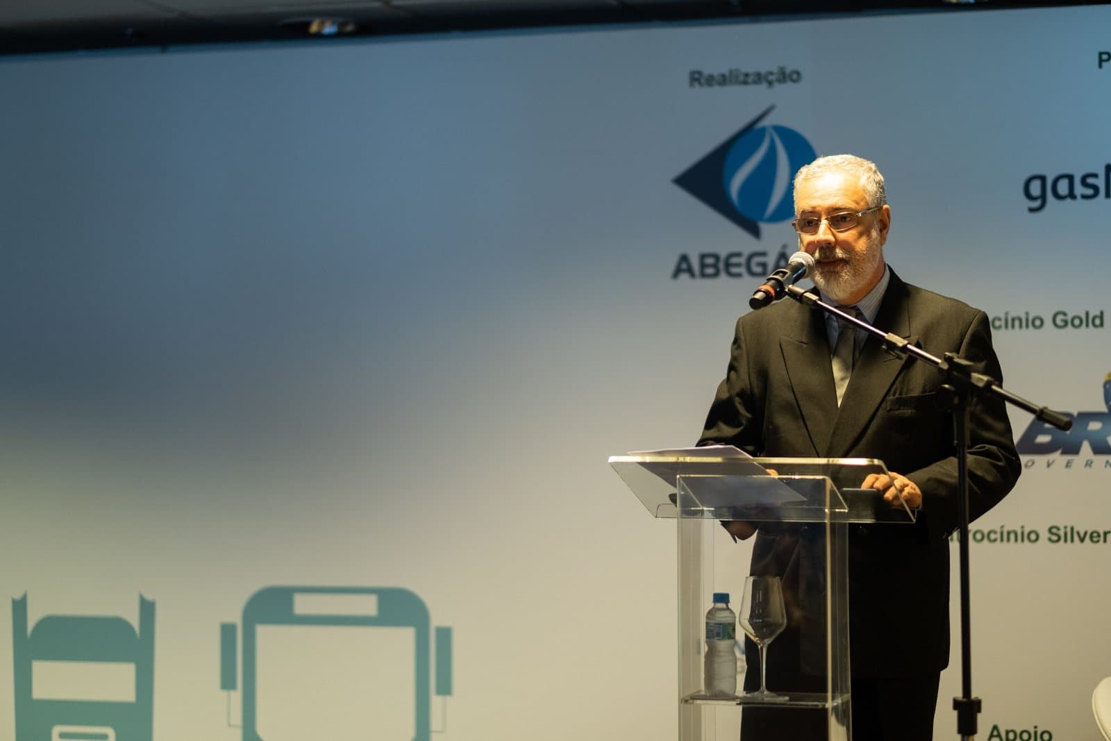 Ruy Jobim de terno ao microfone, num púlpito de acrílico, com o painel da Abegás ao fundo