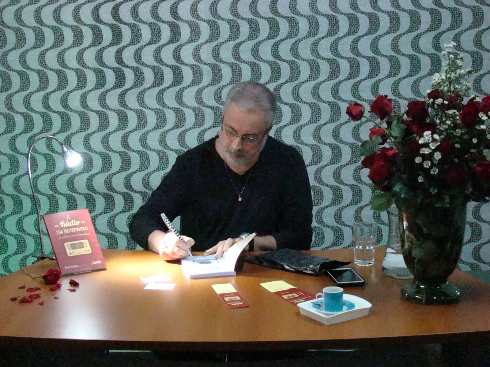 Ruy Jobim autografando exemplares do livro numa mesa com flores, diante de um painel com as ondas do calçadão