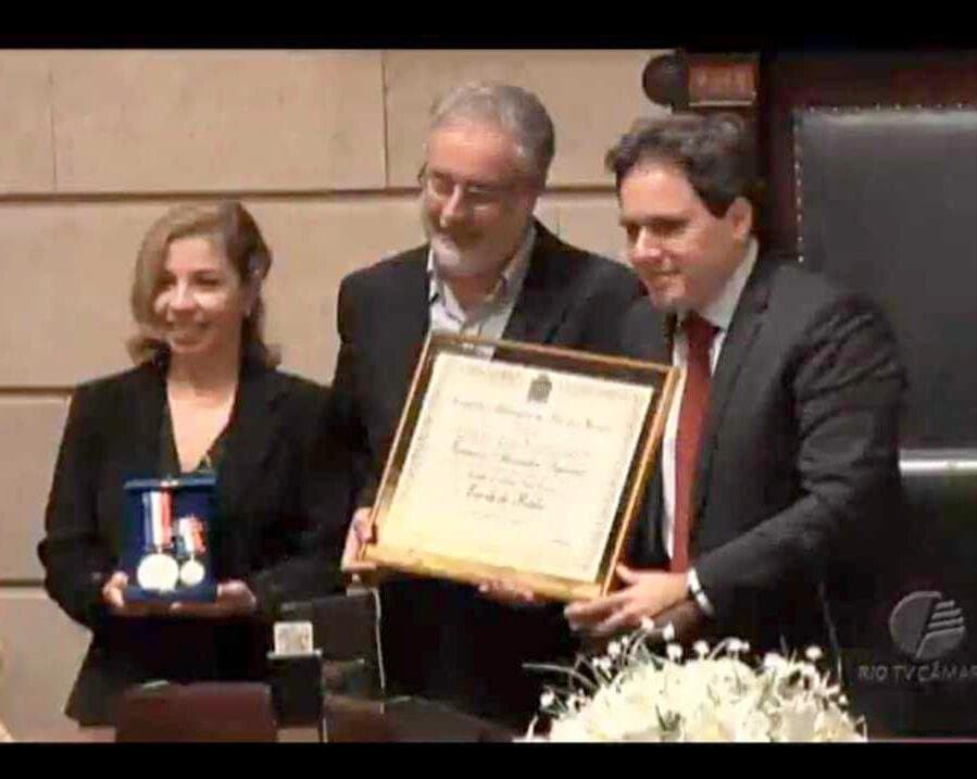 Ruy Jobim segura o diploma da Medalha Pedro Ernesto no plenário da Câmara Municipal do Rio, ao lado de Cristiane, que mostra a medalha